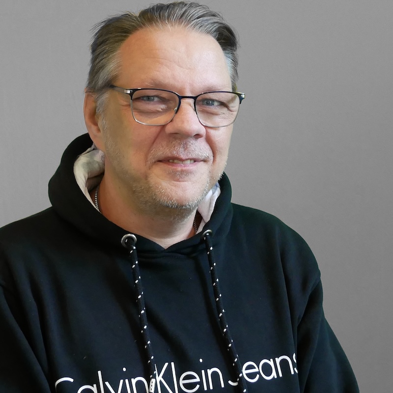 Männlich gelesene Person mit grauen Haaren, Brille in einem schwarzen Calvin Klein Pullover.
