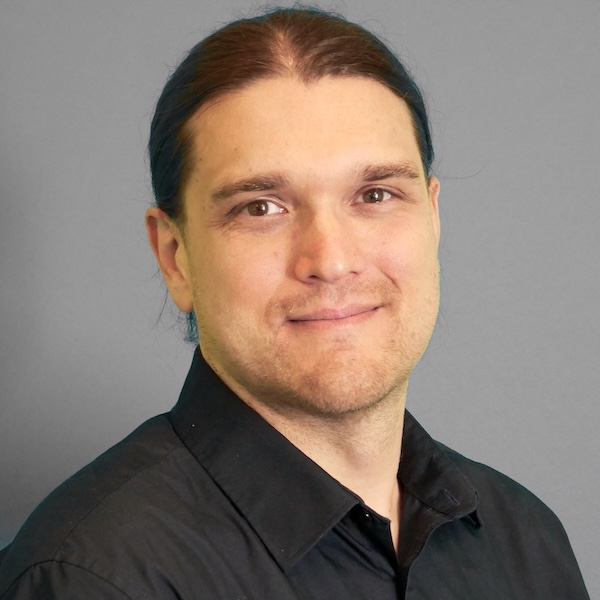 Person mit braun-türkisen Haaren, braunen Augen und einem leichten Bartansatz. Sie trägt ein schwarzes Hemd.