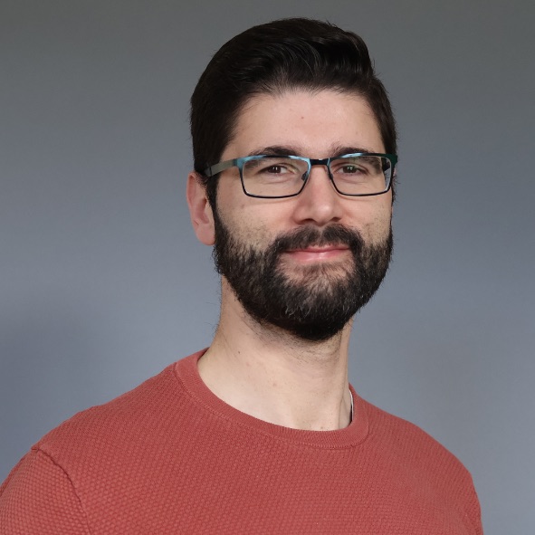 Person mit schwarzen, kurzen Haaren, schwarzem Bart und Brille. Er trägt einen korallenroten Pullover.
